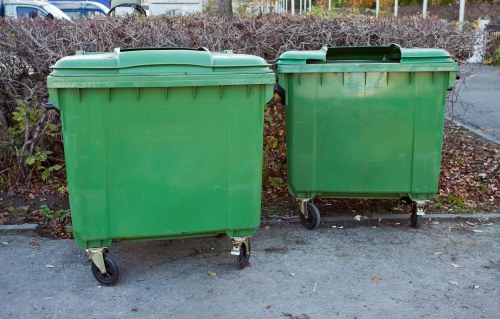 Two green plastic wheelie bins with black wheels and lids are positioned side by side on a paved surface, likely a driveway or loading area. The larger bin on the left has a smooth, matte finish and appears slightly taller and wider than the smaller bin on the right, which has similar features but is one-third smaller in size. Both bins are situated outdoors, with a backdrop of low shrubs or hedges and some blurred fencing or railing behind them. Their surfaces show slight scuff marks and dirt typical of frequent outdoor use, and they are unlabelled, indicating they are containers used for general waste collection. The setting suggests a location where commercial waste removal services, such as those provided by Commercial Waste Removal Haringey, operate, especially within residential or small business areas in or near Haringey. The lighting is natural, with overcast skies casting diffuse light over the scene, emphasizing the utilitarian purpose of these waste containers in a typical urban or suburban environment.