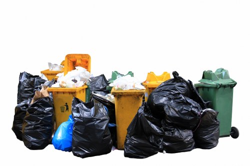 A collection of waste bins and large black rubbish bags positioned outdoors on a paved surface, with some bins in bright yellow and green colours, and the others containing mixed waste materials. The yellow bins have open lids revealing some mixed refuse inside, while the black bags are piled around in front and on top of the bins, indicating a collection or disposal process. The green bin is closed, situated on the right side of the image, and appears to be made of durable plastic with a lid on top. The scene appears to be set on a driveway or paved area, possibly in a residential or commercial environment in Haringey, with a plain white background. The image is related to rubbish removal services, as it shows waste collection containers prepared for disposal or recycling, consistent with the offerings of Commercial Waste Removal Haringey. The lighting suggests natural daylight, with no visible surroundings or background context beyond the waste containers and bags.