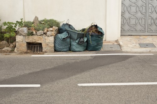 The image displays three large blue/green yard waste collection bags placed on the edge of a paved street next to a white wall with a small garden bed filled with plants and rocks on the left side. The bags are filled with organic debris such as branches, leaves, and garden clippings, and are positioned near a drainage grate embedded in the wall. The bags are situated on the pavement adjacent to the curb, with a narrow strip of asphalt visible in the foreground. The scene appears to be in an urban or suburban area within Haringey, where a waste removal service like Commercial Waste Removal Haringey might collect garden waste and general rubbish. The background includes a section of a decorative metal gate to the right, and the lighting suggests daytime with natural light illuminating the scene evenly. This visual context aligns with rubbish collection and waste management activities in residential or small commercial areas in Haringey.
