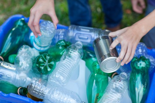 Close-up image of a person's hands sorting through plastic waste, with one hand holding a clear plastic bottle and the other placing a tin can into a bin or collection area, which is filled with various discarded plastic bottles in green, clear, and blue tones. The background suggests an outdoor setting, possibly a garden or park, with green grass visible. The scene emphasizes waste separation and recycling practices commonly associated with rubbish removal services in Haringey. The plastic bottles have varied textures, with some appearing crinkled and others smooth, highlighting different types of plastic materials. The Blue plastic bottles are on the left side, with the person's hands and waste materials arranged centrally and towards the right, capturing a moment of sorting or disposal activity. This visual underlines the importance of proper rubbish management and recycling efforts facilitated by companies like Commercial Waste Removal Haringey.