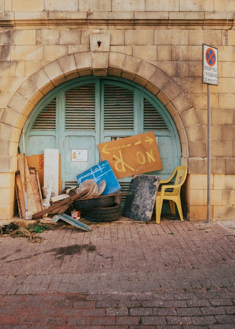 A collection of mixed rubbish is gathered against a large, arched, mint green wooden door set within a yellow brick wall. The waste includes several wooden planks, some leaning against the left side o